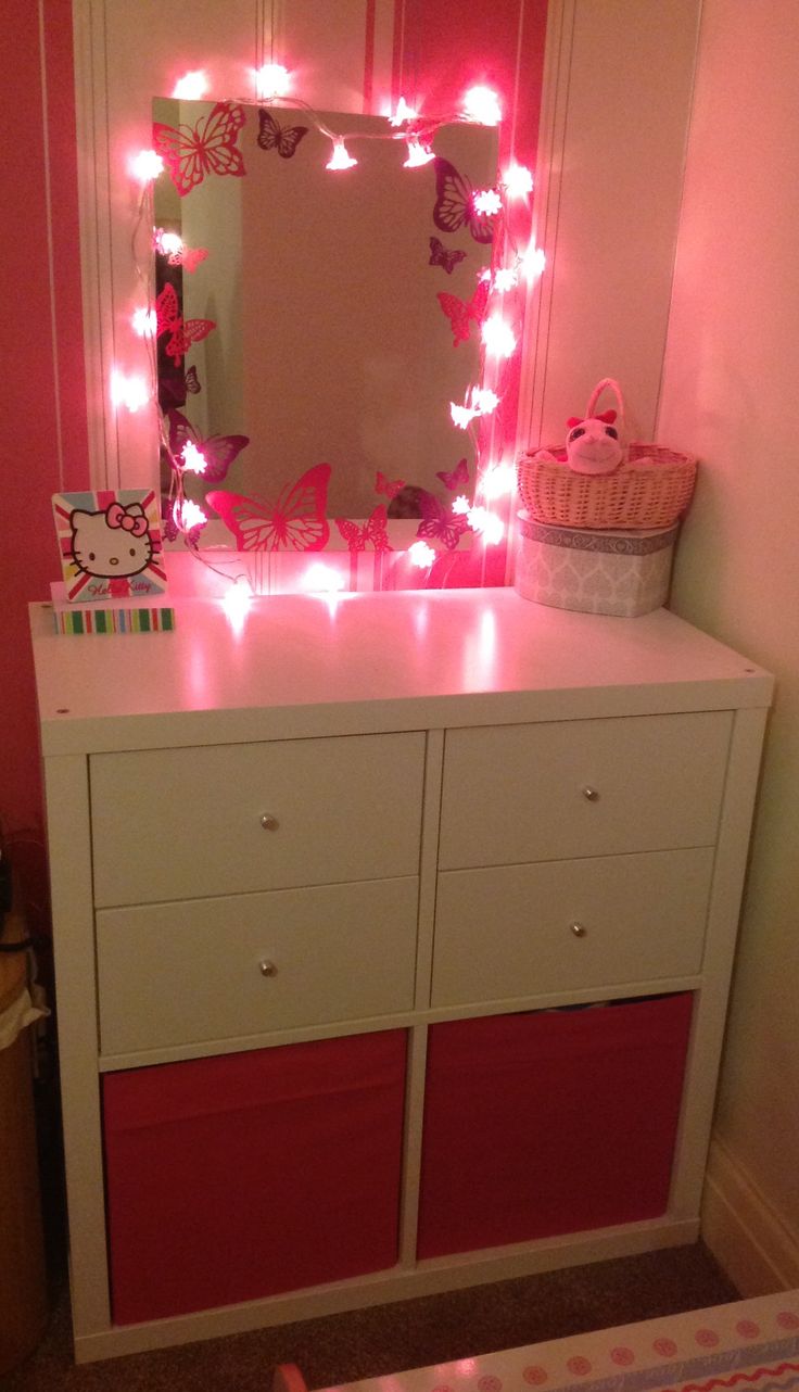 a white dresser topped with a pink mirror and lights on it's sideboard