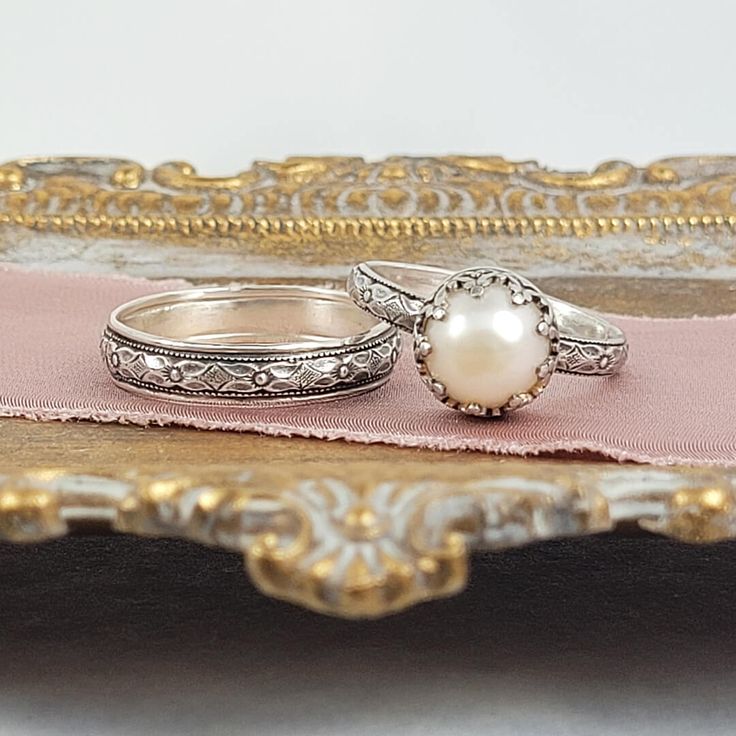 Timeless and classic, this vintage inspired pearl engagement ring and floral wedding band set are reminiscent of the Edwardian era when fine metalwork was designed with intricate details and delicate settings that were fashioned to resemble the look of fine lace.The vintage style Edwardian-inspired pearl ring is handcrafted from recycled sterling silver where a lightly oxidized floral band is set with a luminous 8-8.5mm cultured creamy white freshwater pearl in a filigree heart-crown setting.The Wedding Jewelry With Milgrain Detail In Round Band, Silver Wedding Jewelry With Historical Design, Elegant Milgrain Jewelry For Wedding, Wedding Jewelry With Milgrain In Round Band, Milgrain Round Jewelry For Wedding, Elegant Milgrain Wedding Jewelry, Heirloom Pearl Ring With Intricate Design, Silver Historical Design Jewelry For Wedding, Silver Jewelry With Historical Design For Wedding