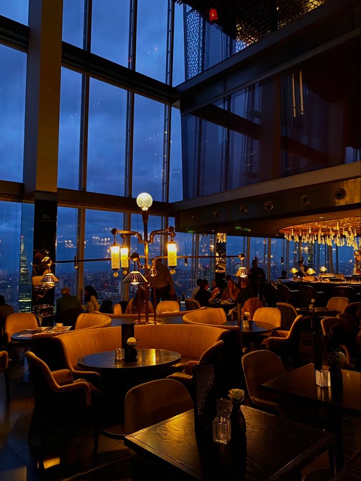 people are sitting at tables in a restaurant with large windows overlooking the city lights and skyscrapers