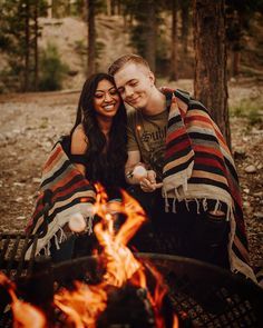 a man and woman wrapped in blankets sitting next to a campfire