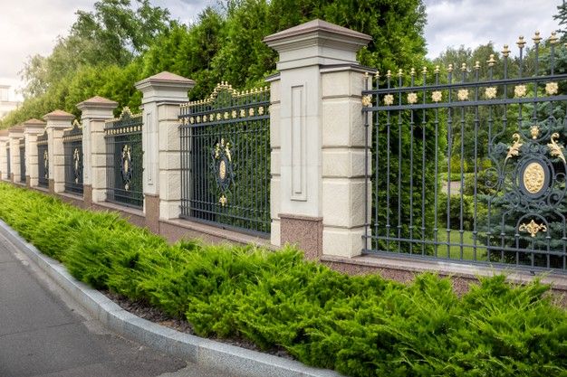 an iron fence with trees and bushes along the side of it, in front of a street