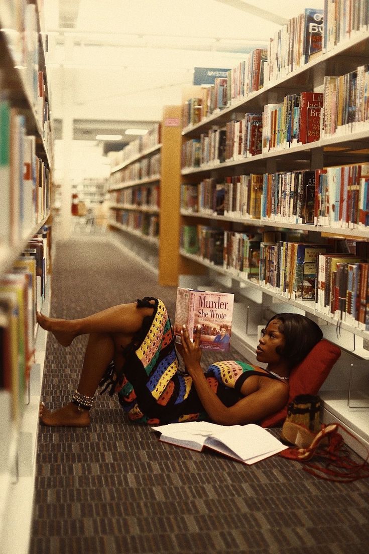 a woman sitting on the floor reading a book in a library with bookshelves behind her