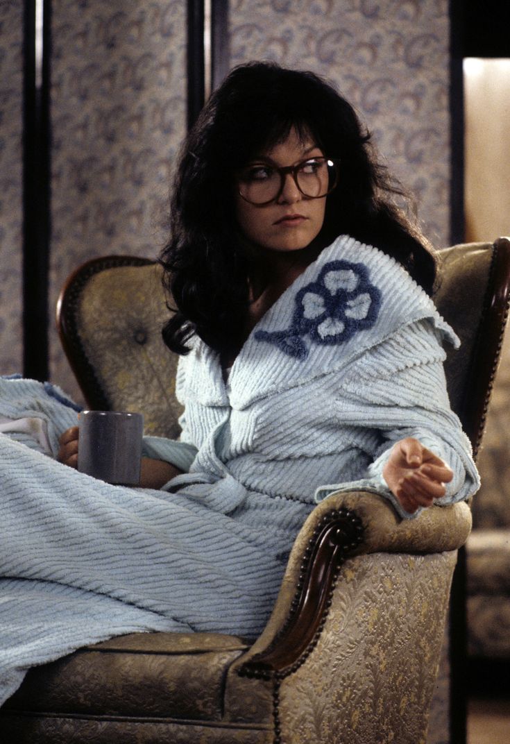 a woman sitting in a chair holding a coffee cup and looking at the camera while wrapped in a blanket