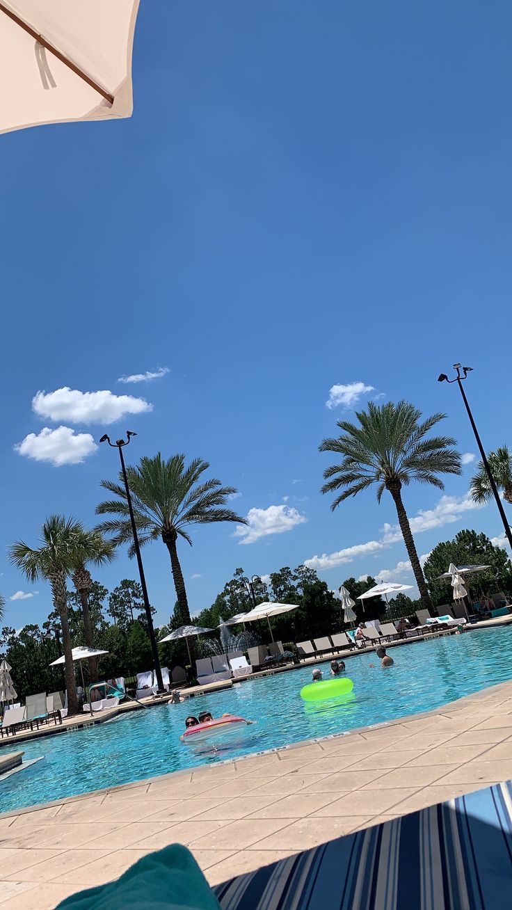 people are swimming in an outdoor pool surrounded by palm trees and umbrellas on a sunny day