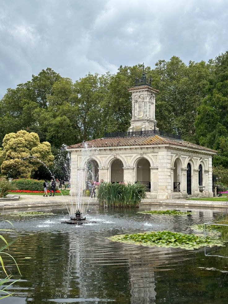 a small fountain in the middle of a park with people walking around and trees surrounding it