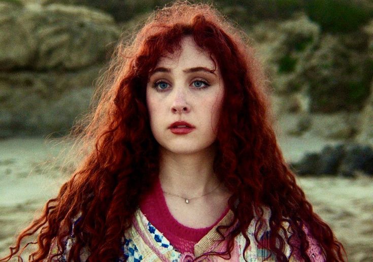 a woman with long red hair standing in front of some rocks and looking at the camera