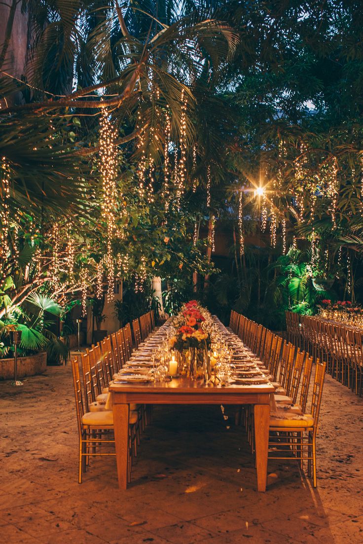 an outdoor dinner table set up with candles and flowers in the center, surrounded by greenery