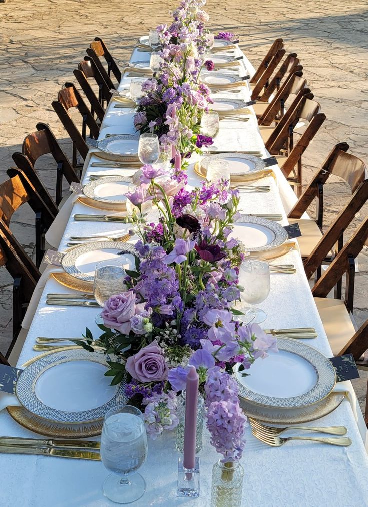 a long table is set up with purple flowers and place settings for the dinner guests