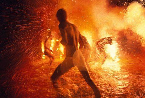 a man running in front of a fire with lots of flames behind him and another person standing next to him