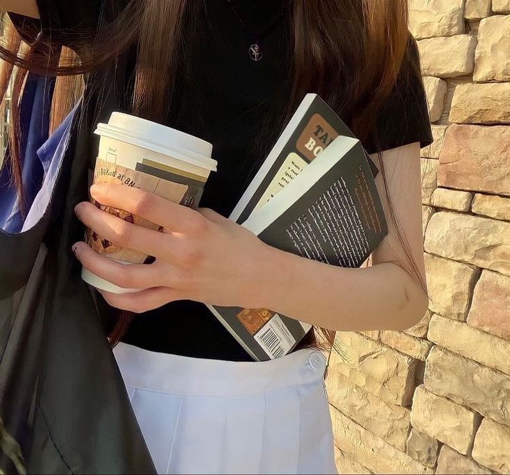 a woman holding a cup of coffee and some books in her hands while standing next to a brick wall