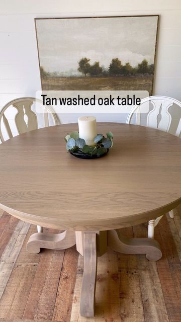 a wooden table with two white chairs and a candle on it, in front of a painting that reads tan washed oak table