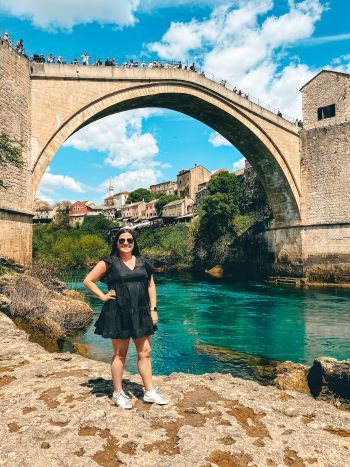 a woman standing on the edge of a river with an old bridge in the background