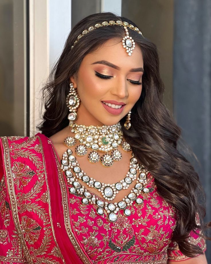 a woman wearing a pink and gold bridal outfit with jewelry on her neck, in front of a window