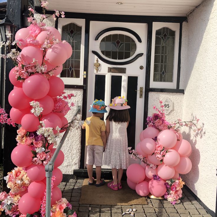 two children standing in front of a house with pink balloons and flowers on the door