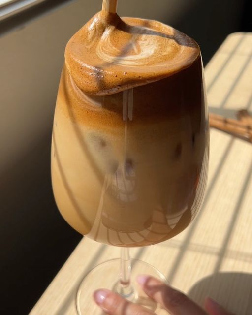 a hand holding a wine glass filled with liquid on top of a wooden table next to a window