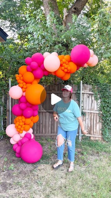 a woman standing in front of a pink and orange arch with balloons attached to it