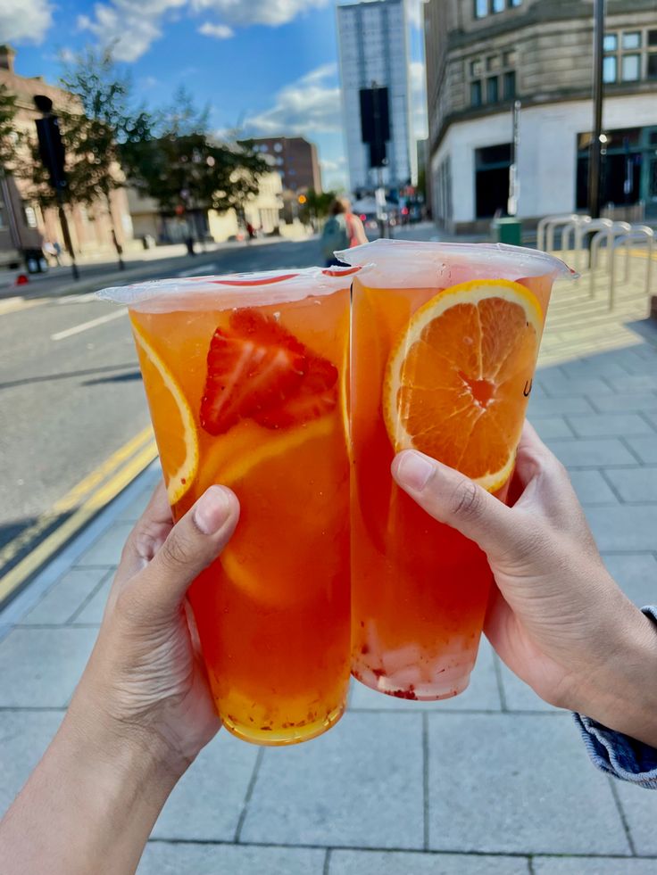 two people hold up glasses with orange juice and strawberries in them on the sidewalk