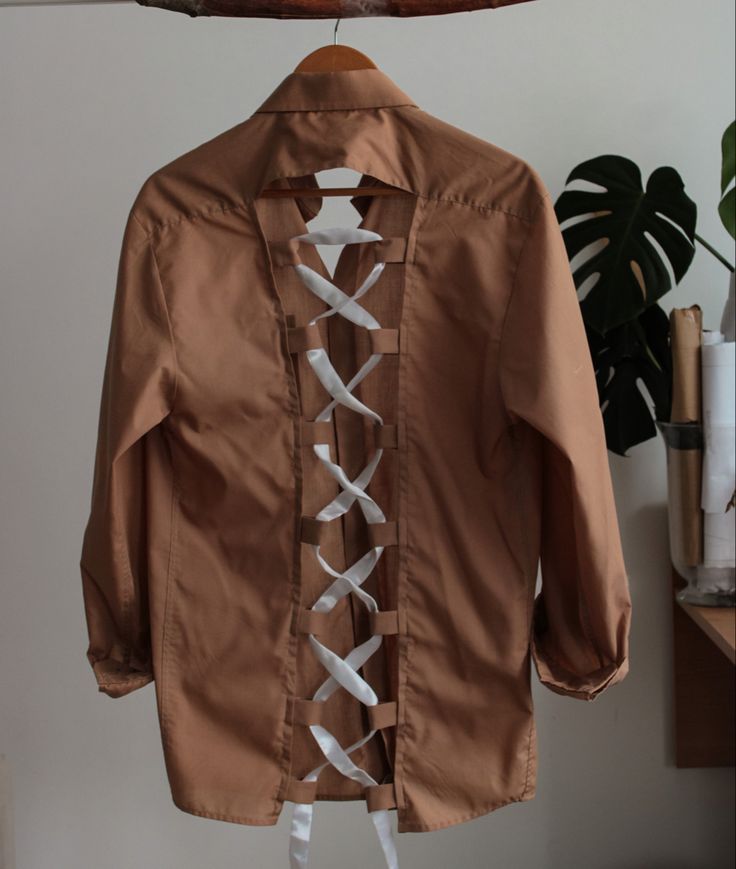 a brown jacket with white laces hanging on a clothes hanger next to a potted plant