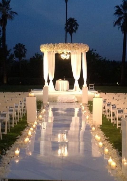 an outdoor wedding setup with white chairs and lit candles on the aisle at night, surrounded by palm trees