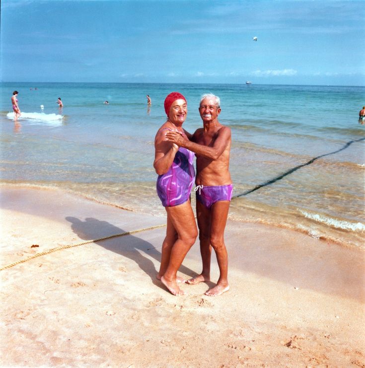 two older people in bathing suits standing on the beach with their arms around each other