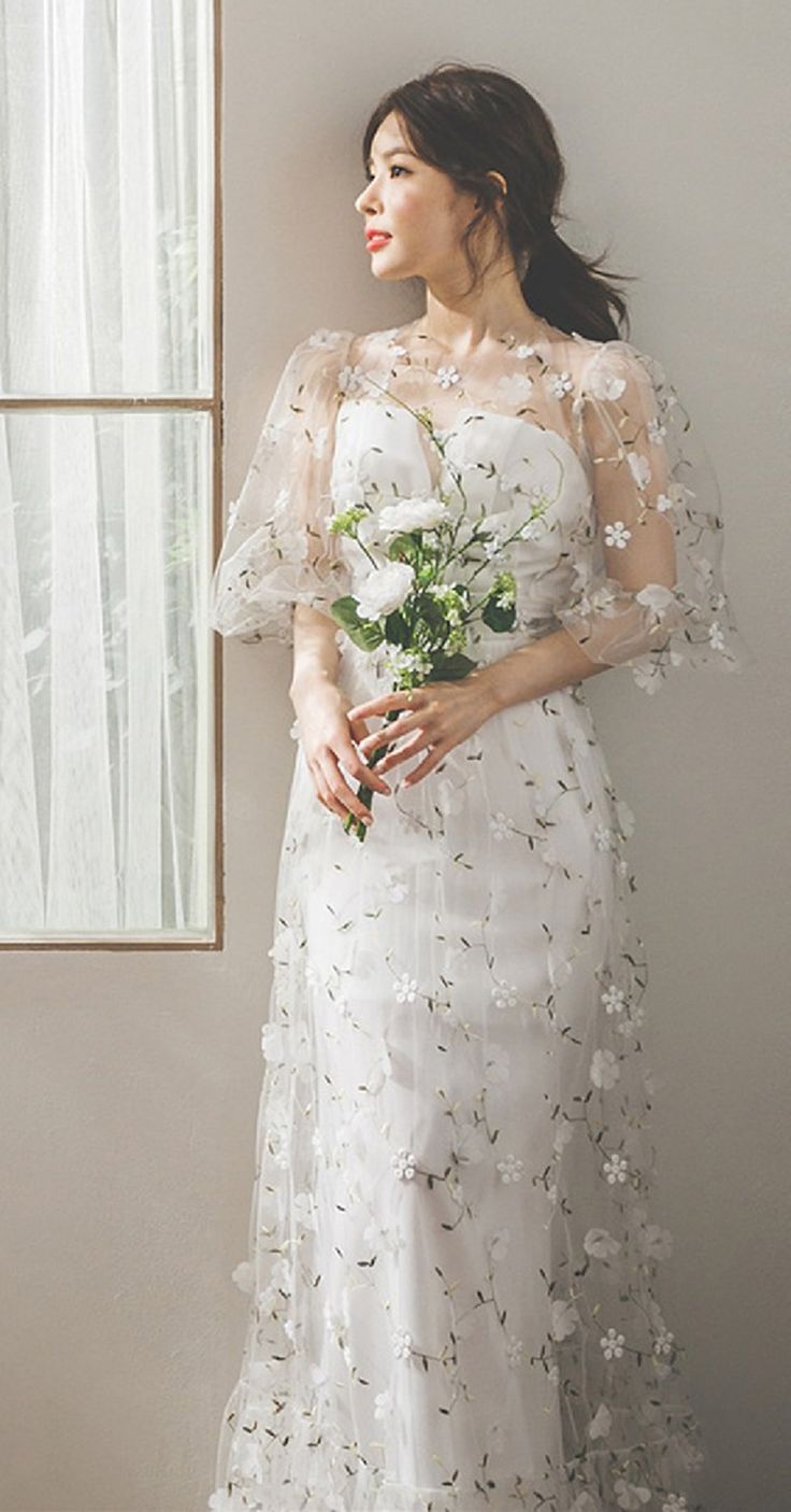 a woman standing in front of a window wearing a white dress with flowers on it