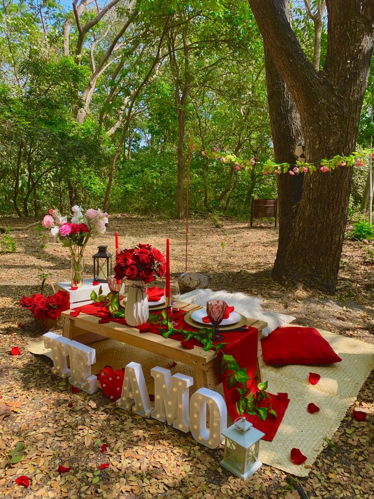 a picnic table set up in the woods with red and white flowers, candles and napkins