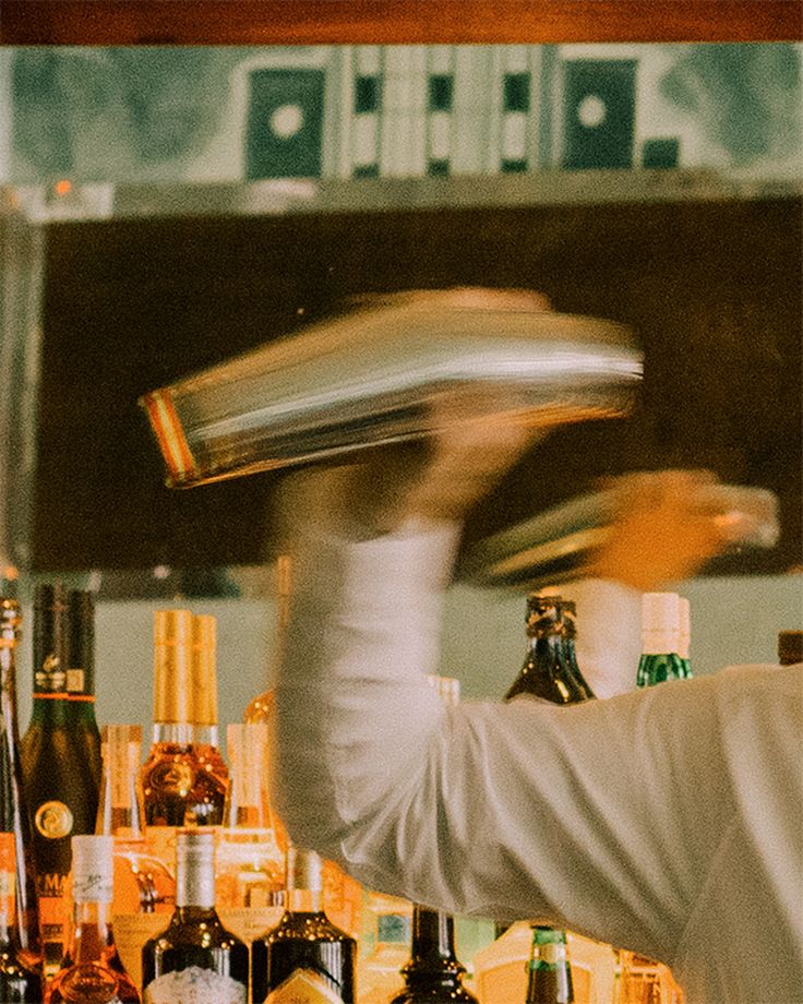 a man in a white coat is making a drink at a bar with liquor bottles behind him