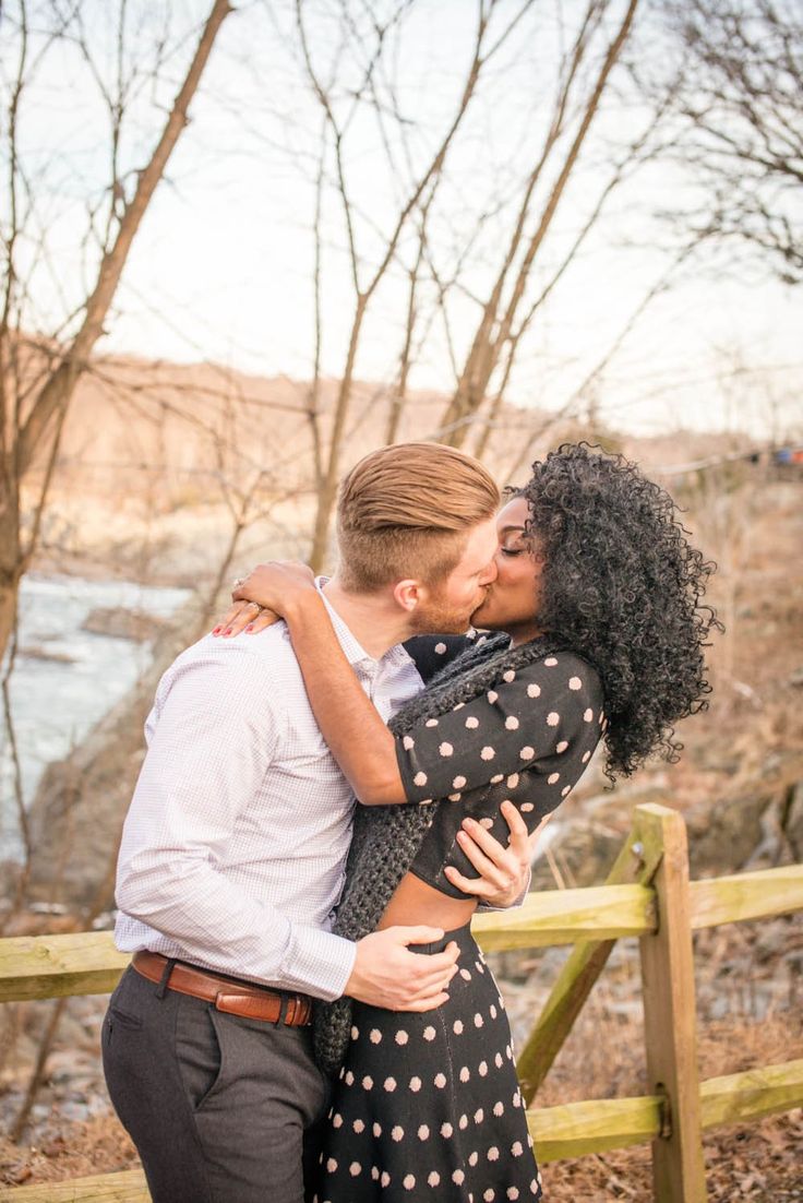 a man and woman are kissing in front of a wooden fence with water behind them