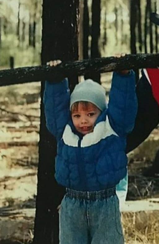 a young boy holding onto a wooden fence in the woods with his hands up above his head