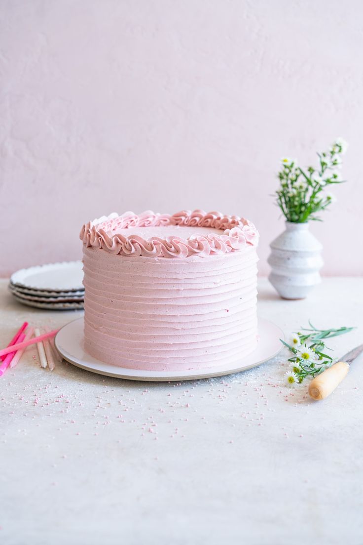 a pink frosted cake sitting on top of a white plate next to a vase