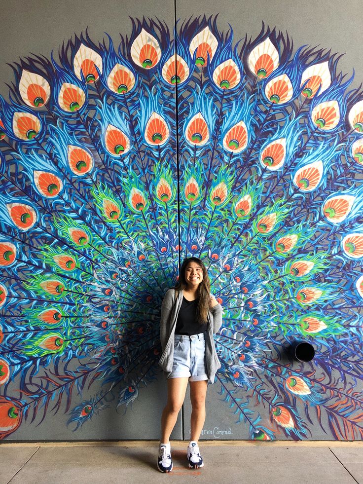 a woman standing in front of a colorful peacock mural