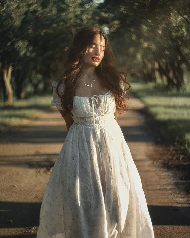 a woman in a white dress is standing on a dirt road with trees behind her