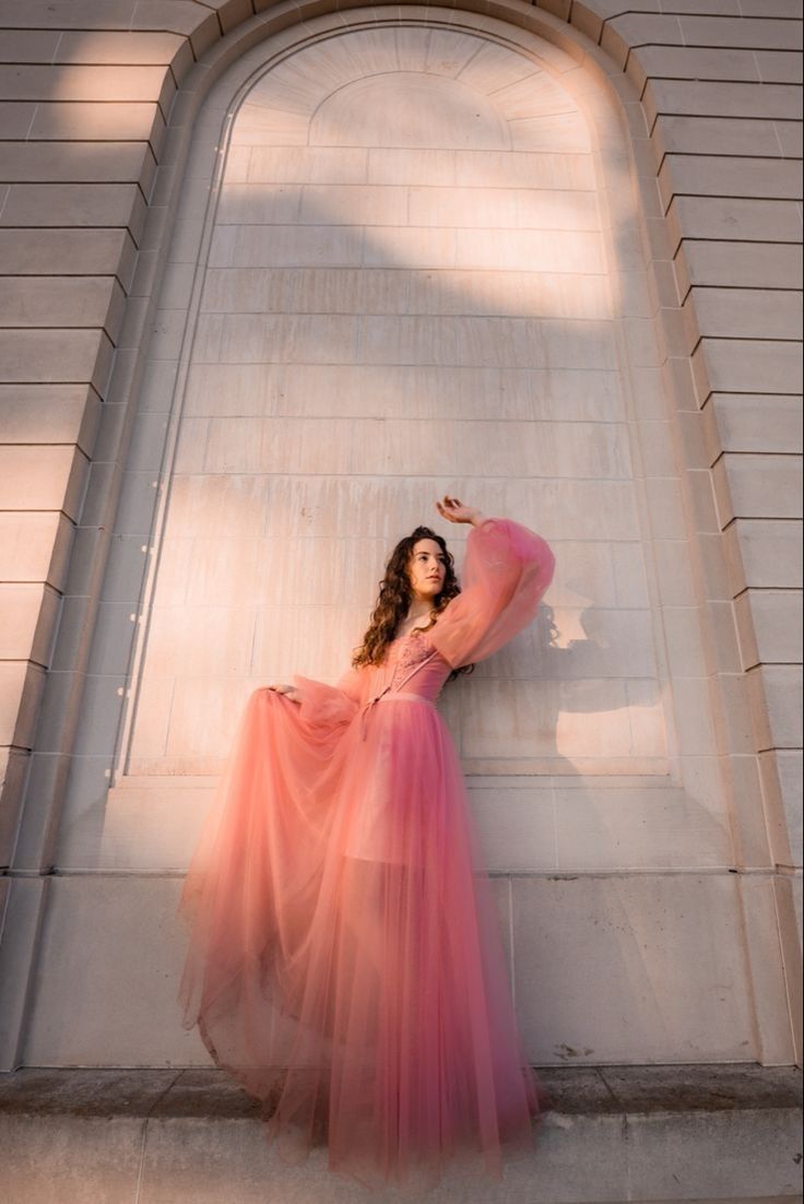 a woman in a long pink dress leaning against a wall with her hands on her hips