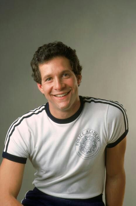 a man in a white shirt and black shorts smiling at the camera with his hands on his hips