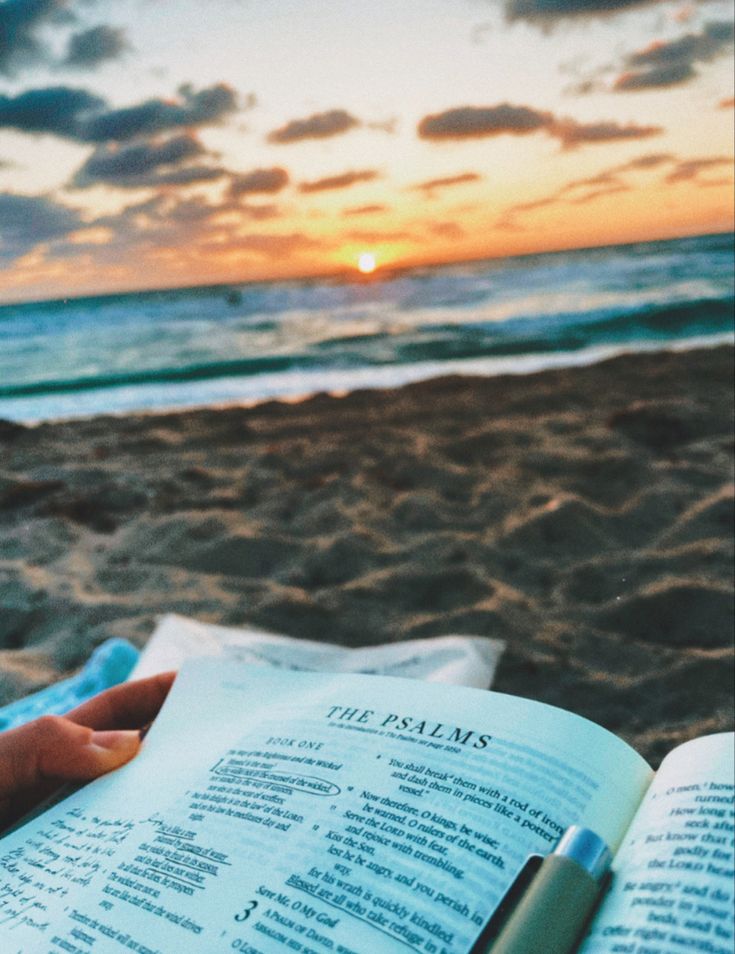 a person is reading a book on the beach at sunset or sunrise with their hand resting on an open bible