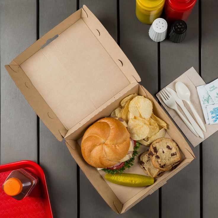 an open box containing a sandwich, chips and pickles on a table with other food items