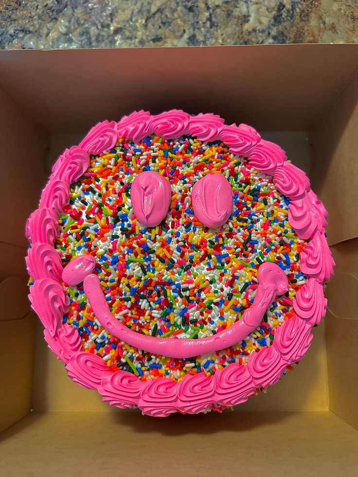 a pink cake with sprinkles and smiley face on top in a box