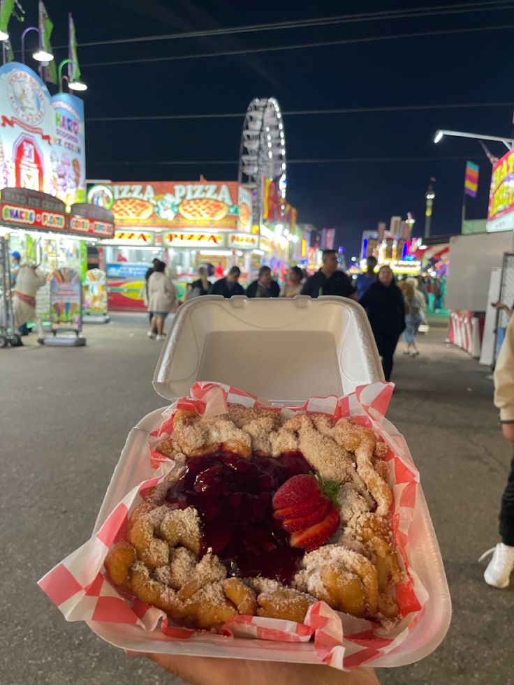 a person holding up a pastry with strawberries on it in front of carnival rides