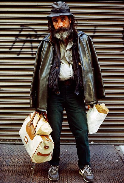 a man with long hair and beard wearing a leather jacket holding two bags while standing in front of a garage door