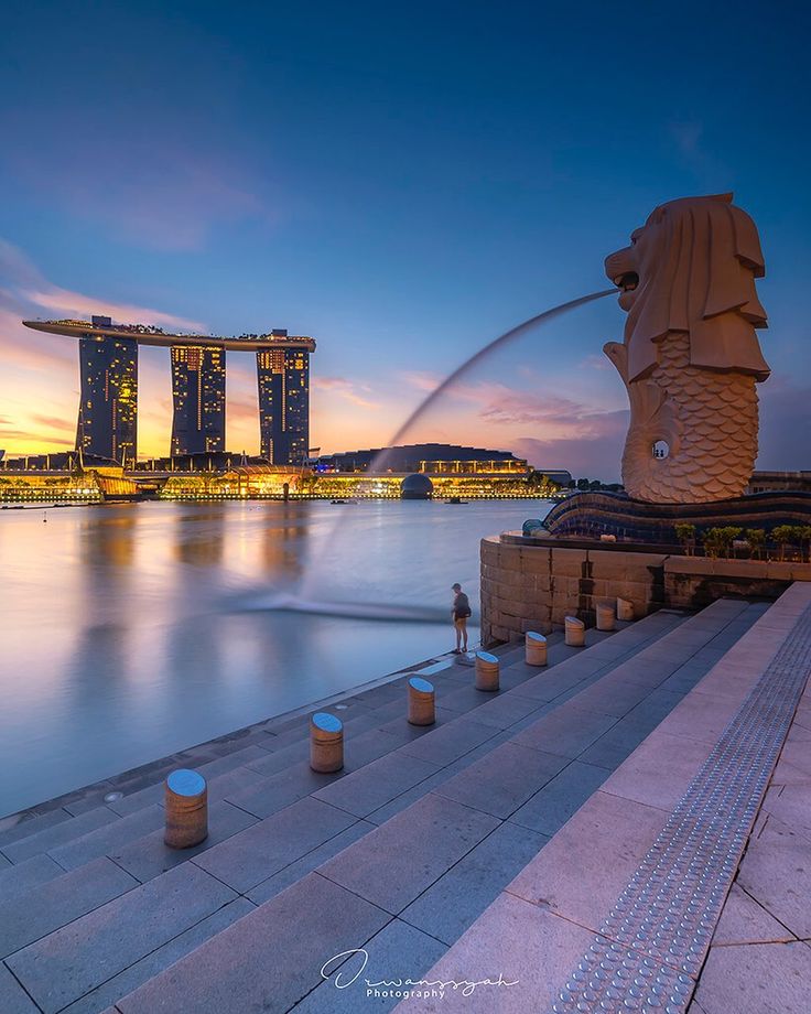 the statue is in front of an ocean and cityscape at dusk with water spouting from its mouth