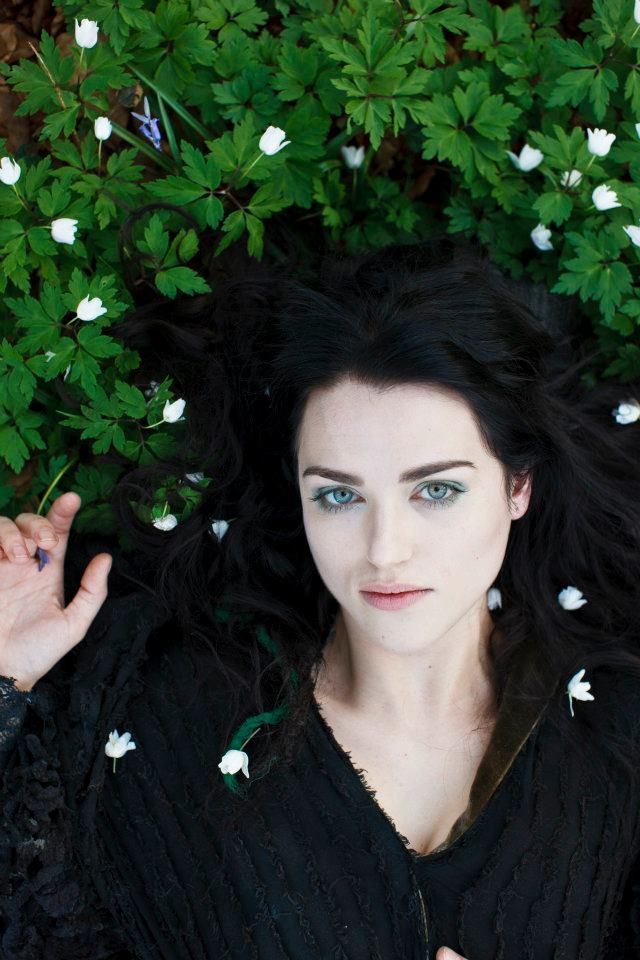 a woman with long black hair and blue eyes is laying on the ground surrounded by flowers