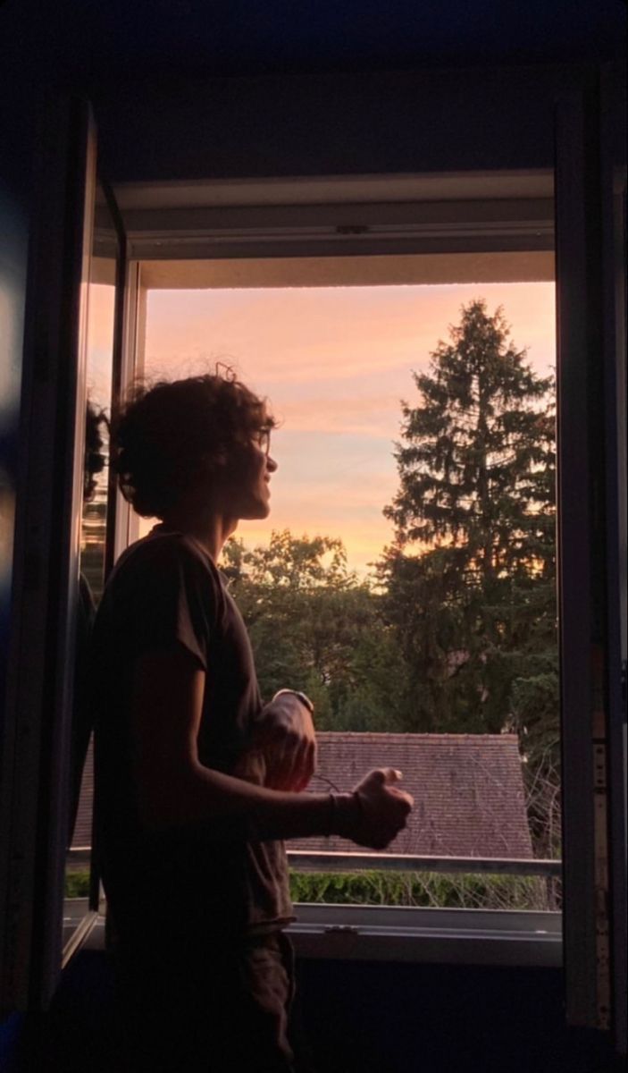 a person standing in front of a window holding a cup and looking out at the sunset