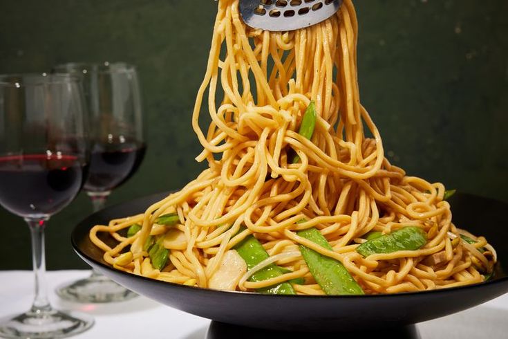 a plate with noodles and vegetables on it next to two glasses of red wine in the background