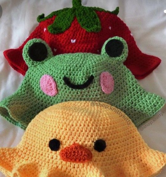 three crocheted hats with faces on them, one has a strawberry and the other is a frog