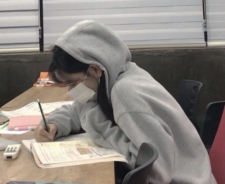 a girl wearing a hoodie writing in a notebook with a cell phone on the table