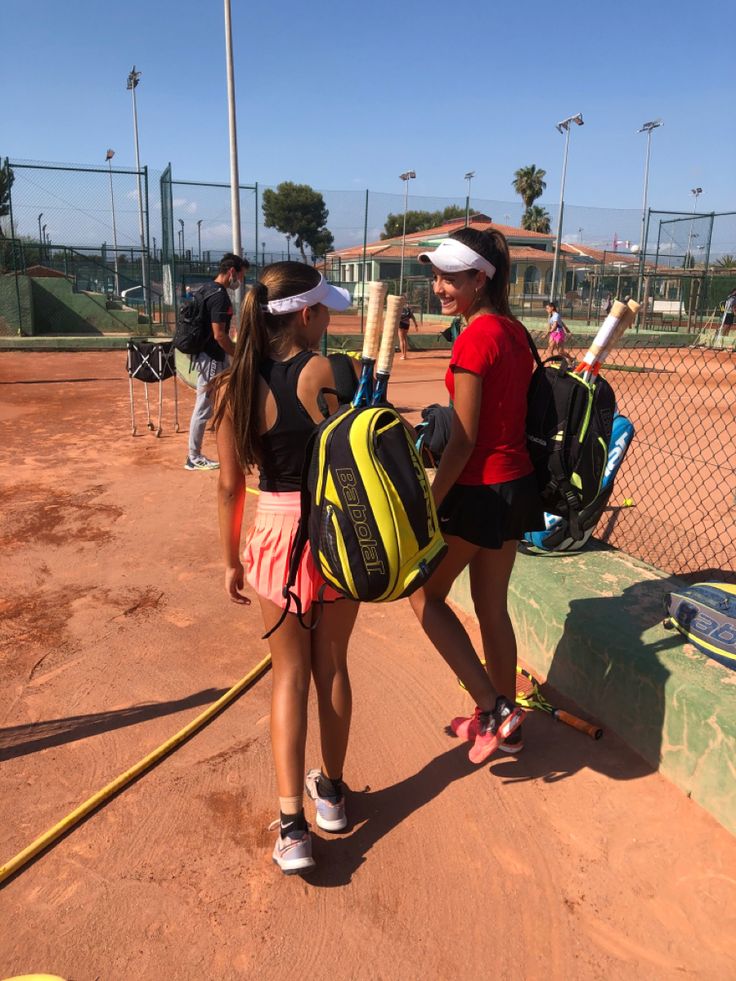 two girls with backpacks and baseball bats on a dirt field in front of a fence