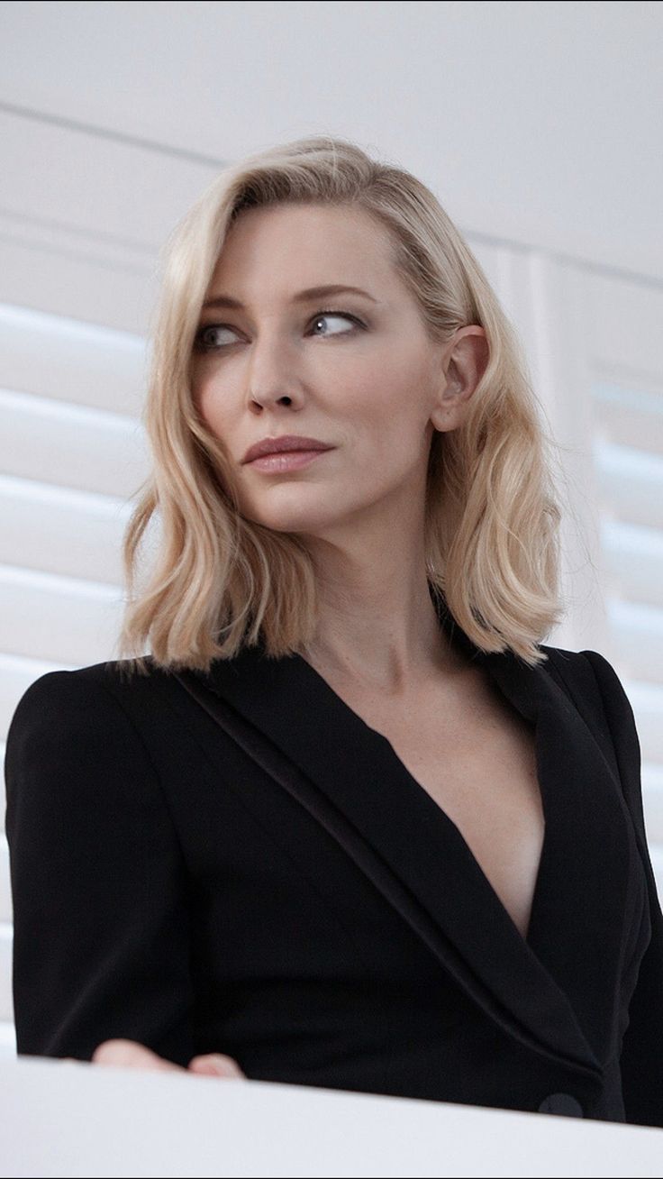 a woman with blonde hair wearing a black dress and looking off into the distance while standing in front of white shutters