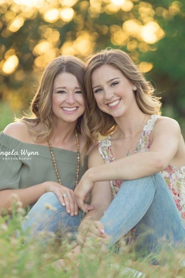 two women are sitting in the grass and posing for a photo with their arms around each other