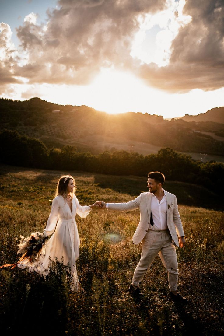 a bride and groom holding hands while walking through a field with the sun setting behind them