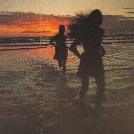 two people are standing in the water at sunset on the beach with their hair blowing in the wind